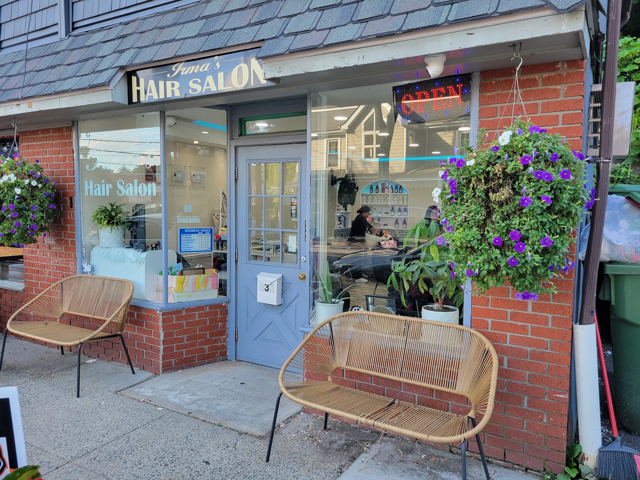 The storefront of Irma's Hair Salon on Grand Street: red brick, a blue door, two rattan benches and hanging baskets of purple petunias